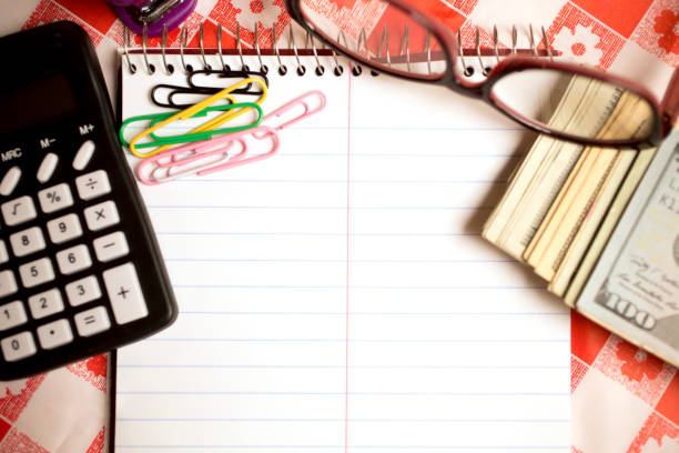 Kitchen table with notepad, calculator, cash currency, eye glasses, paper clips and stapler.  Calculating about refinancing decisions.