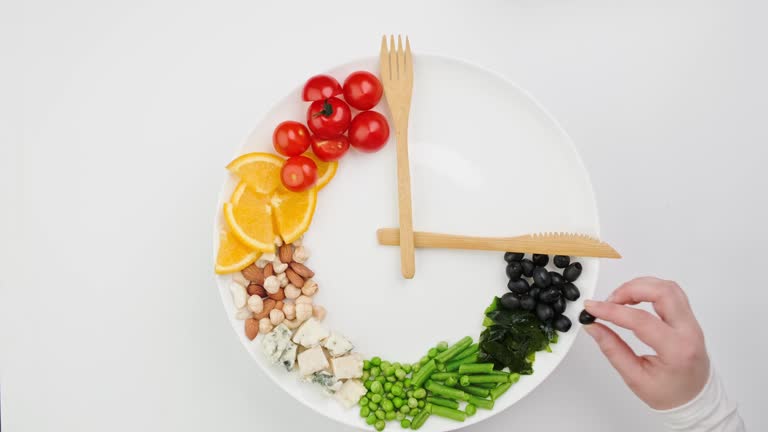 Colorful food and cutlery arranged in the form of a clock on a plate. Hand takes an olive. Intermittent fasting, diet, weight loss, lunch time concept.