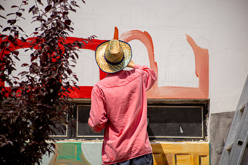 One male artist painting wall mural outdoors with bigger brush. He is making colorful strokes.