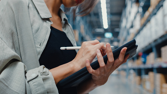 Attractive young Asia businesswoman manager looking for goods using digital tablet checking inventory levels standing in retail shopping center. Distribution, Logistics, Packages ready for shipment.