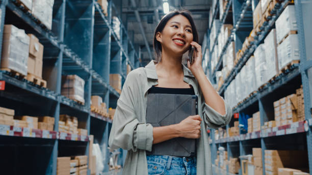 joven empresaria de asia vendedora en línea hablando confirmando pedidos de clientes en el teléfono que sostiene la tableta digital de pie en el centro comercial minorista. - industry asian ethnicity asia manager fotografías e imágenes de stock