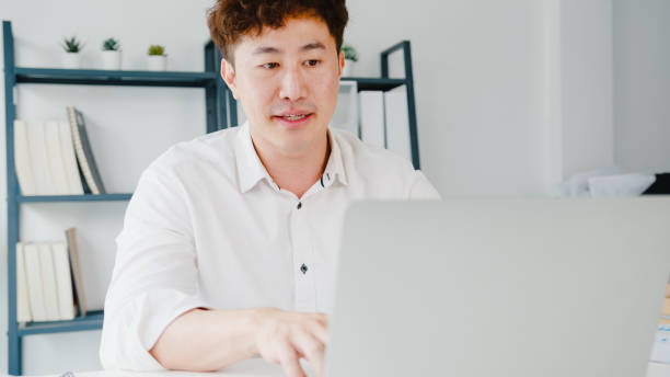 istock Young Asia businessman using laptop talk to colleagues about plan in video call while smart working from home at living room. 1320998643