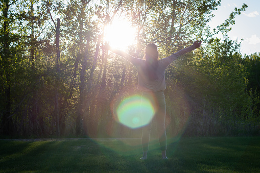 Young woman facing landscape away from camera with arms outstretched embracing nature with sun and sunflare and copy space, trees and sky in background