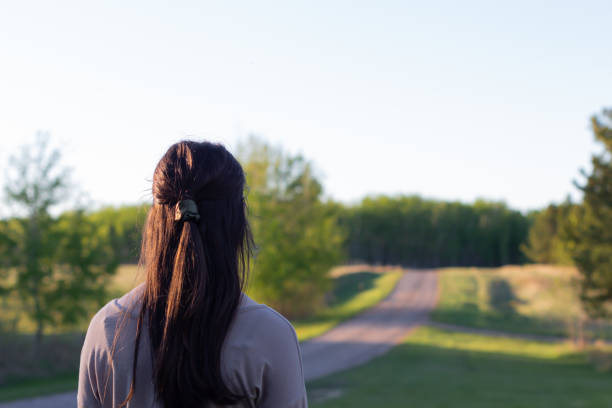 young woman with dark brown hair looking away from camera up a rural road into the distance with trees and green grass in evening sun with copy space