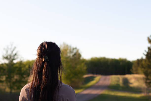 young woman with dark brown hair looking away from camera up a rural road into the distance with trees and green grass in evening sun with copy space