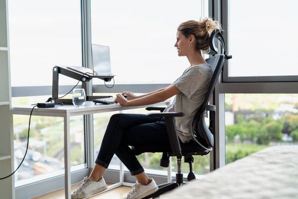 Side view of mid adult woman sitting on desk while doing home office during daytime Smiling woman working from home while sitting at her desk in the morning office chair stock pictures, royalty-free photos & images