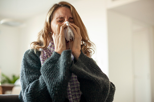 Mid adult woman wearing a scarf and sweater while blowing her nose in living room during daytime