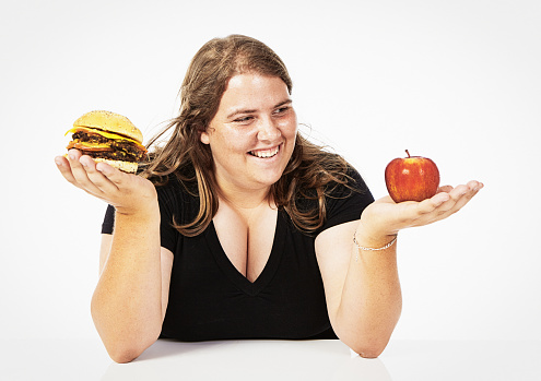 Young woman smiles at an apple she's holding, about to choose it over the burger in her other hand.