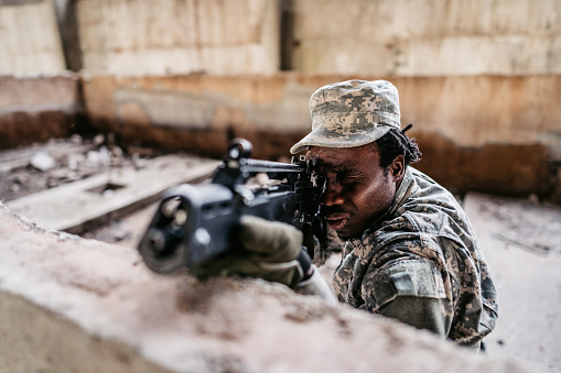 A young male soldier looks into the gun sight in order to aim his military assault rifle.