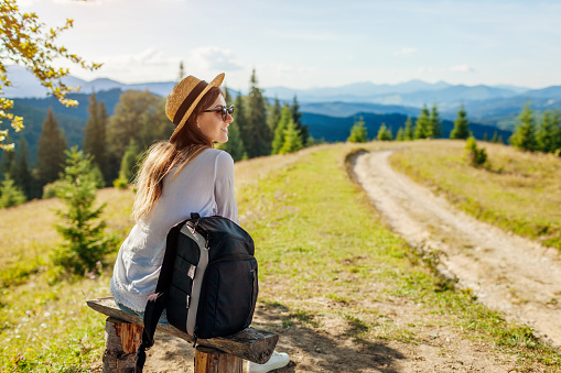 Trip to Carpathian mountains. Woman tourist hiking and relaxing admiring landscape sitting on bench under tree with backpack. Traveling around spring Ukraine