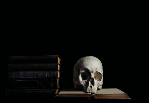 Photo of witchcraft human skull laying on old books and stacked books on black background.