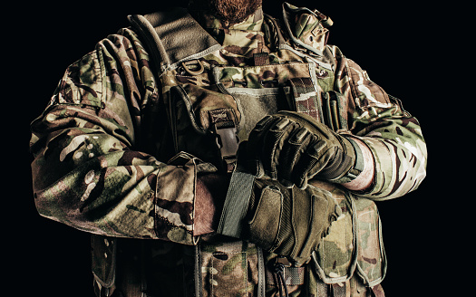 Photo of soldier in level 3 camouflaged armored vest putting on tactical gloves on black background.