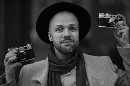 A Russian man walks around Moscow, takes pictures of the world around him with a film camera. Fashionable man in a hat and coat. Blurred background. Black and white portrait.