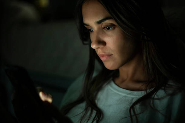 Portrait of young woman looking at screen while holding mobile phone in the dark stock photo