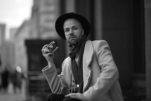 A Russian man walks around Moscow, takes pictures of the world around him with a film camera. Fashionable man in a hat and coat. Blurred background. Black and white portrait.