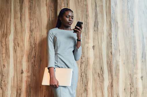 Portrait of successful African-American businesswoman wearing mask and using smartphone while standing in sunlight by wood background, copy space