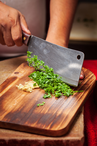 Close-up of hands chopping fresh green coriander or parsley leaves on a wooden cutting board.