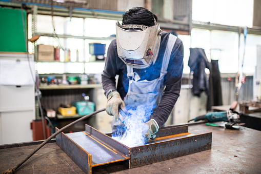 Man in protective helmet/mask welding a steel girder. 
From the typical day in the life series of a Japanese man working at a steel factory in Japan.