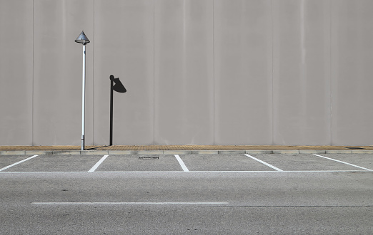 Gray concrete wall with a streetlight and its shadow. Sidewalk and asphalt road with parking in front. Background for copy space.