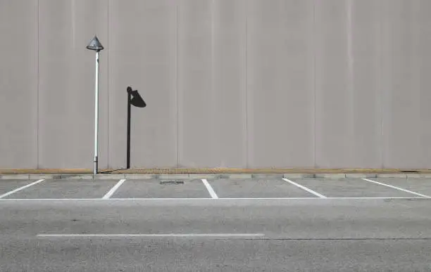 Gray concrete wall with a streetlight and its shadow. Sidewalk and asphalt road with parking in front. Background for copy space.