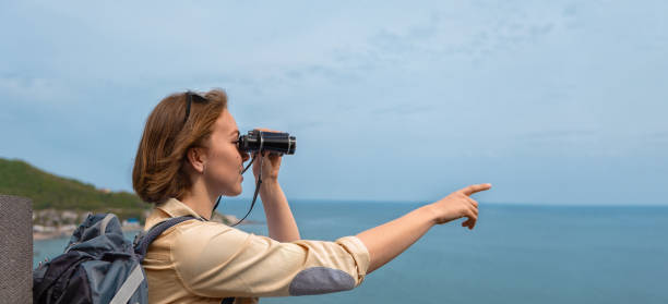 istock Woman tourist with a backpack looking through binoculars and points his hand against the sea and the blue sky.with copy space 1321077244