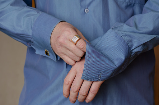 Close-up of hands buttoning a cufflink on the sleeve of a blue shirt. A middle-aged adult man with a gold ring on his index finger. Person holding hands in middle section of body. Lifestyle. Indoors.