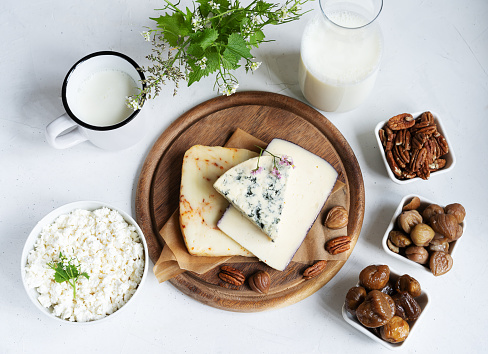 Different kind of cheese sliced on the wooden cutting board, surrounded be chestnuts. pecan nits, dry figs, milk jar, kefir and cottage cheese. Flat lay view