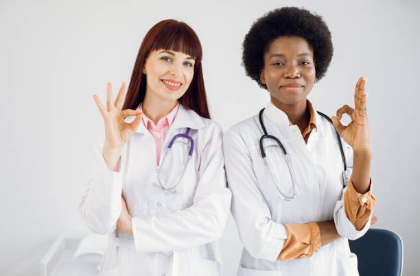 istock Okay, cool, alright, approval sign gesture, making circle with fingers. Two pleasant smiling cheerful female multiracial doctors in lab coats and with stethoscopes around neck, showing ok sign. 1321090721