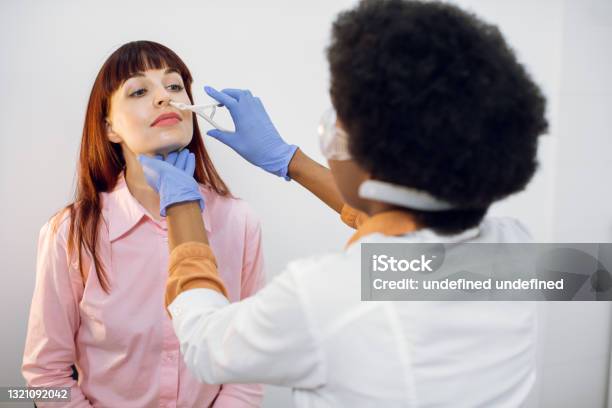 Back View Of Female Afro Doctor Ent Doing Nose Examination Of Her Patient Young Caucasian Lady Sitting On The Chair And Looking At Her Doctor During Nasak Check Up Nose Deseases Concept Stock Photo - Download Image Now