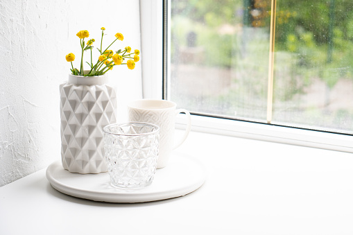 White home decoration with yellow flowers, ceramic vase and glass on tray, summer interior decor objects close-up