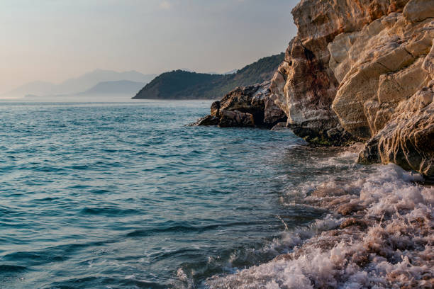Beautiful landscape - beach in sunset sunlight - waving sea water, rocks and cliffs, blue sky and mountains on the horizon.