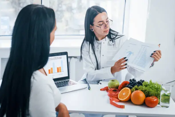 Female nutritionist with laptop gives consultation to patient indoors in the office.