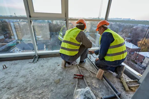 Photo of Two workers in protective clothing and safety helmets installing plastic pipes using soldering iron on balcony of flat of building under construction