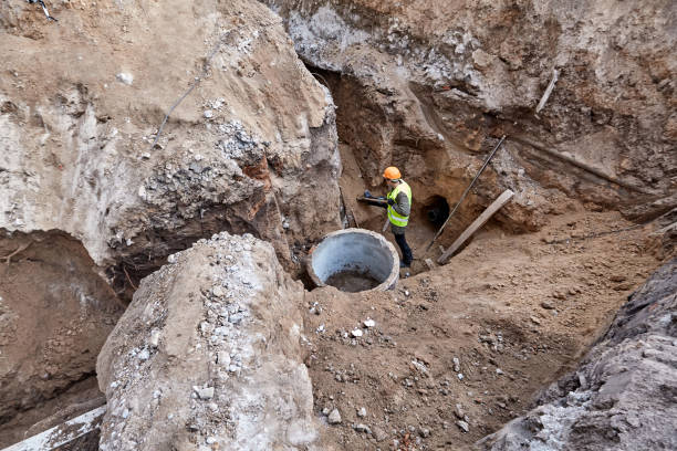 Worker is digging out a trench with a shovel in an excavation pit to install communications to a new high-rise building under construction