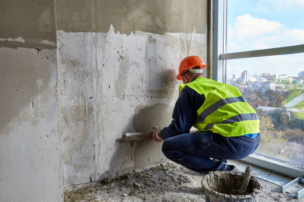 istock Skilled worker in working clothes and orange hard hat is putting putty on the wall, using spatula. Flat of a building under construction 1321114929