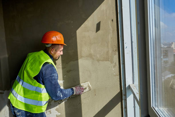 istock Worker in workwear and safety helmet is putting putty on wall, using spatula. Apartment of building under construction with panoramic windows 1321114959