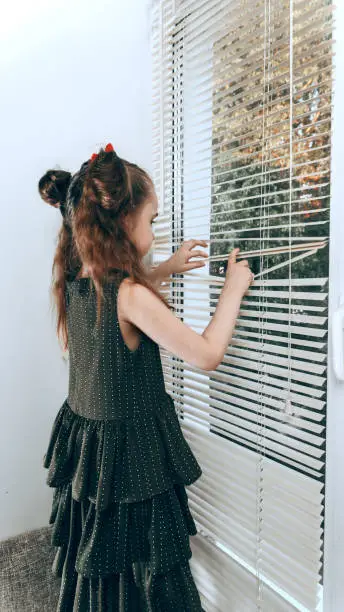 Photo of A baby girl in a green handmade dress, looks through the blinds out the window. A girl with curvy ponytails. Selective soft focus