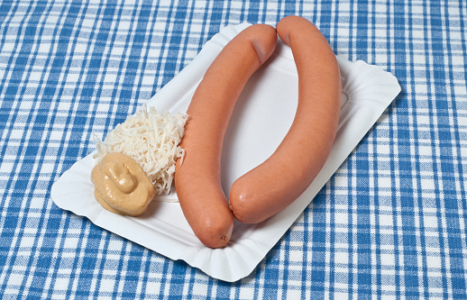 A pair of pair of Frankfurter sausages with mustard and kren (horse radish) on a white cardboard plate.  The base is a blue cloth napkin.