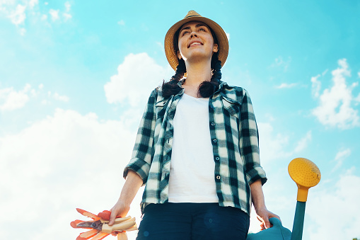 Woman with gloves and a watering can in her hands standing in the garden. Bottom view. Sky on the background. Copy space.