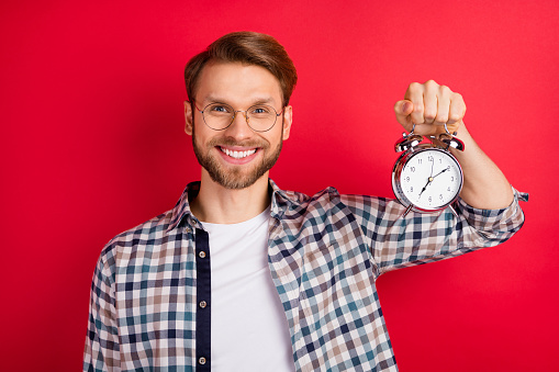 Photo of funny charming young guy wear checkered shirt glasses holding retro clock isolated red color background.