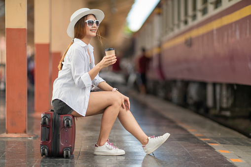 Beautiful Asian traveler wearing hat and glasses holding a cup of coffee while sitting on luggage at the railway station, waiting for departure.