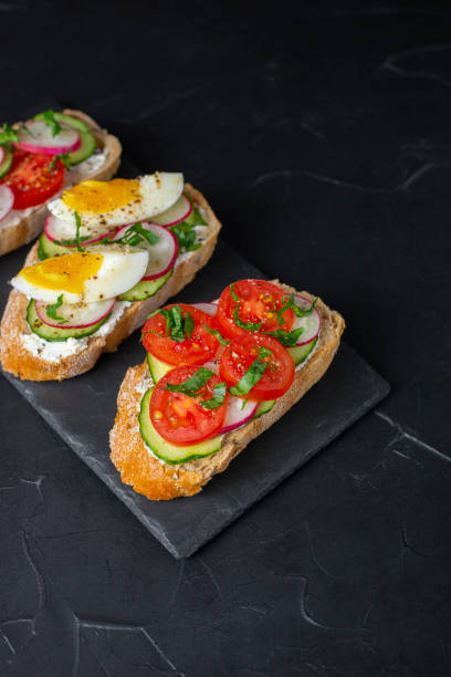 istock Delicious toasted sandwiches white bread with cream cheese, vegetables: cherry tomatoes, cucumbers, eggs, parsley. Healthy vegetarian breakfast idea. Served on black stone board, dark background 1321147675