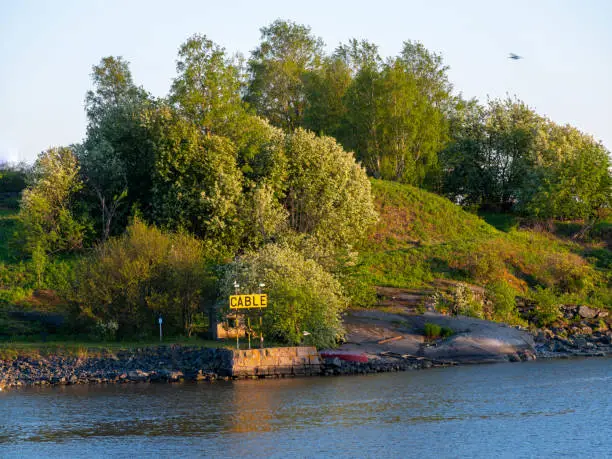 Helsinki / Finland - MAY 29, 2021: Golden sunset casting beautiful light on an island and trees. A warning sign indicating under-the-sea cable route.