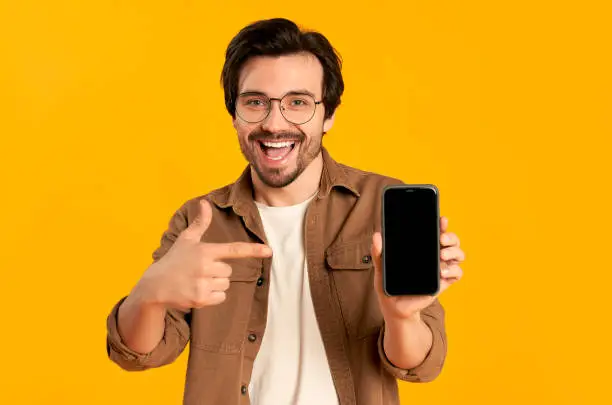 Young bearded man in glasses shows on blank smartphone screen isolated on orange background.