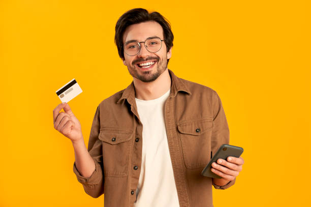 Courier Young bearded man in glasses holds credit card and smartphone isolated on orange background. Online shopping. Online payment. credit card stock pictures, royalty-free photos & images