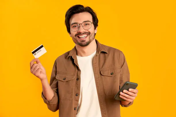 Young bearded man in glasses holds credit card and smartphone isolated on orange background. Online shopping. Online payment.