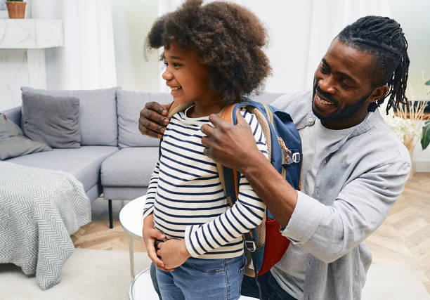 papá afroamericano feliz ayuda a la hija a prepararse para la escuela y le pone una bolsa de la escuela en la espalda - relación humana fotografías e imágenes de stock