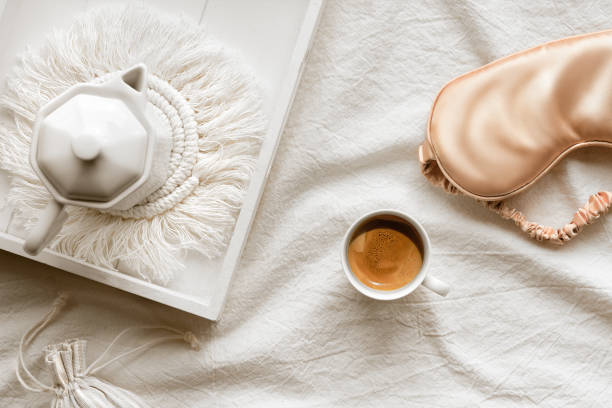 Espresso in hand, tray with white ceramic espresso coffee maker on trendy macrame pad. Sleep mask on crumpled bed sheets. Cup of espresso. Good morning Simple minimal top view