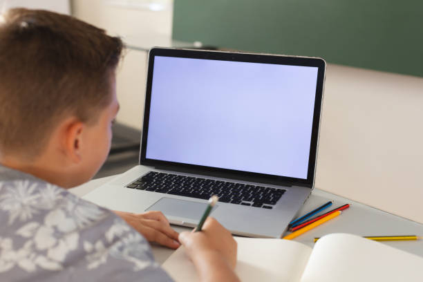 istock Caucasian boy sitting at a desk in classroom writing and using laptop, with copy space on screen 1321183153