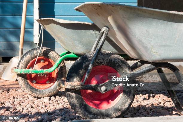 Two Dirty Wheelbarrows With Silver Bodies And Red Wheels Sit On The Gravel Driveway A Building Wall And A Shovel Are Visible In The Background Closeup Background Stock Photo - Download Image Now
